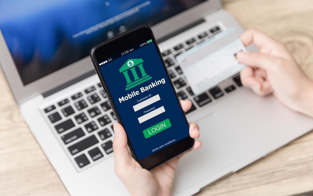 Mobile banking – Enter your customer number and password to log in. A person is holding a smartphone with a login screen and a credit card, with a laptop in the background.