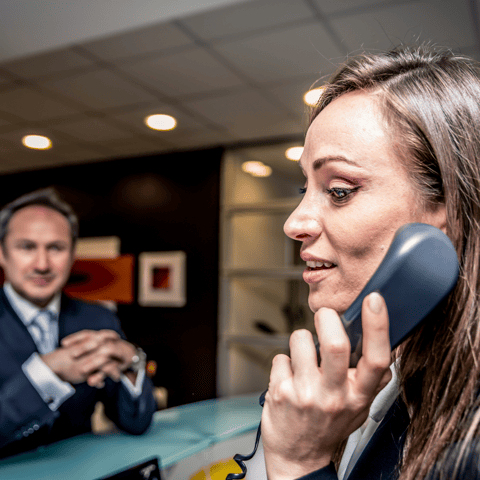 Hotel receptionist answering a customer by IP phone. There is a customer waiting to be attended to at reception.