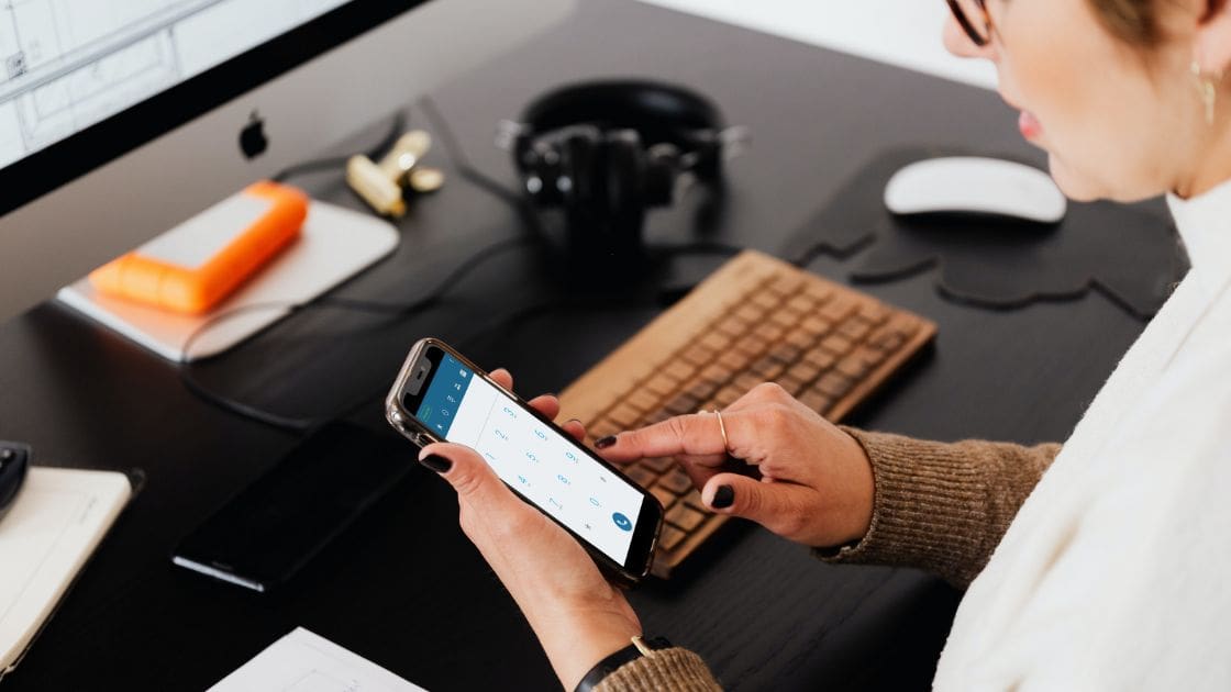 A person is sitting at a desk using a smartphone. On the desk are a computer screen, a keyboard, headphones, and an external hard drive.