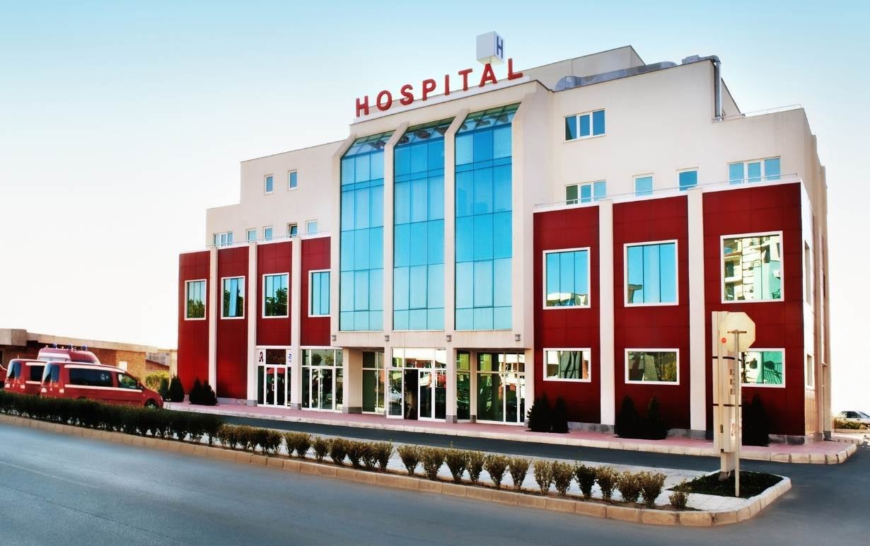 Modern hospital building with a red and white facade, large glass windows, and an "HOSPITAL" sign. Red ambulances are parked outside, with a clear sky and road in the foreground.